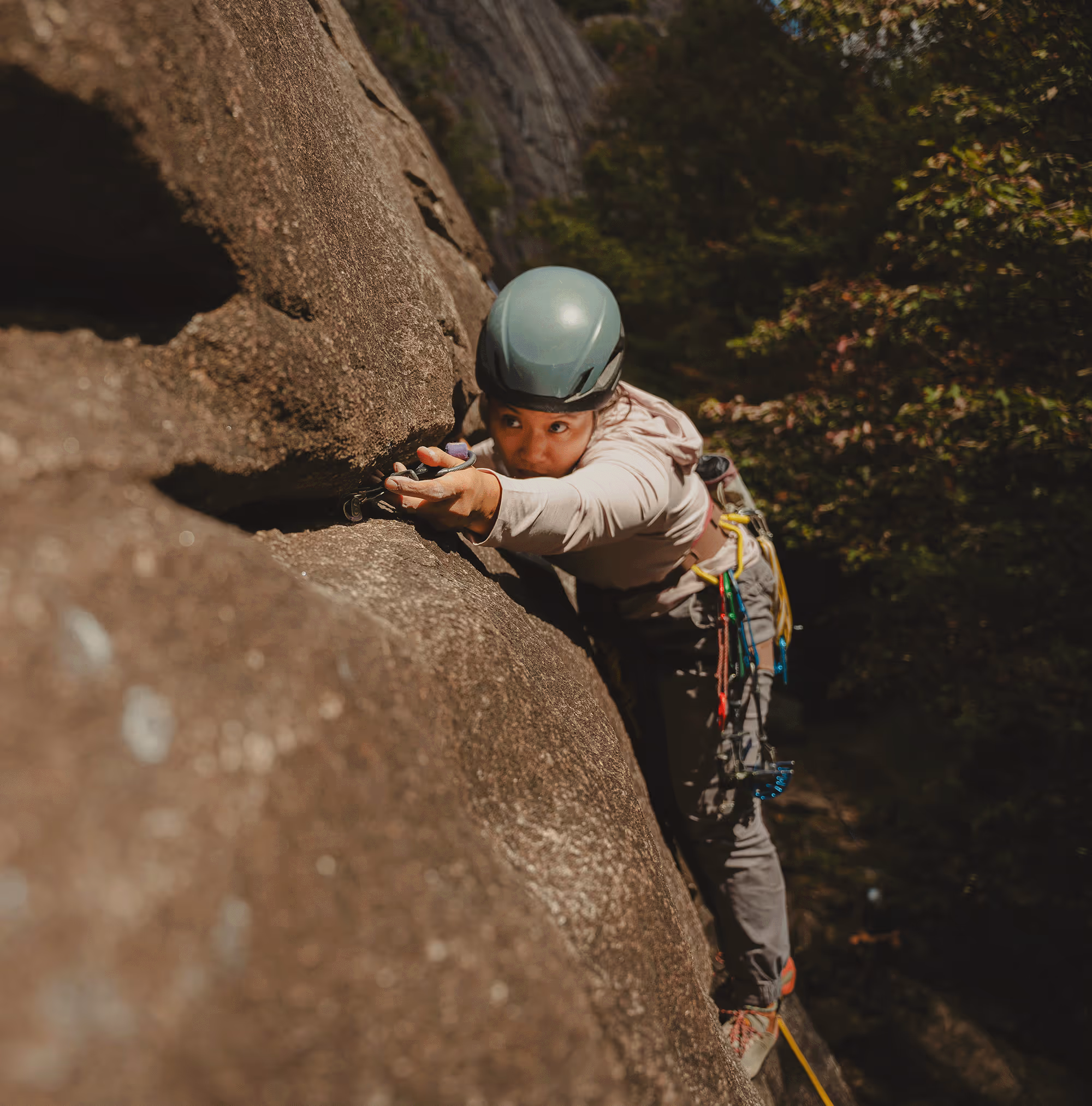 A person in climbing gear scales a steep rock face, reaching for a handhold.