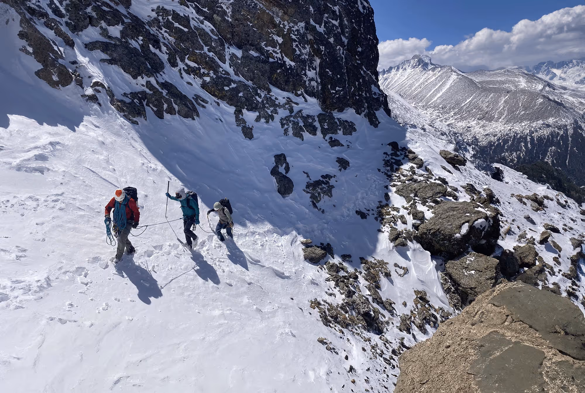 Three climbers hike up a snowy mountain slope, using ropes for safety.