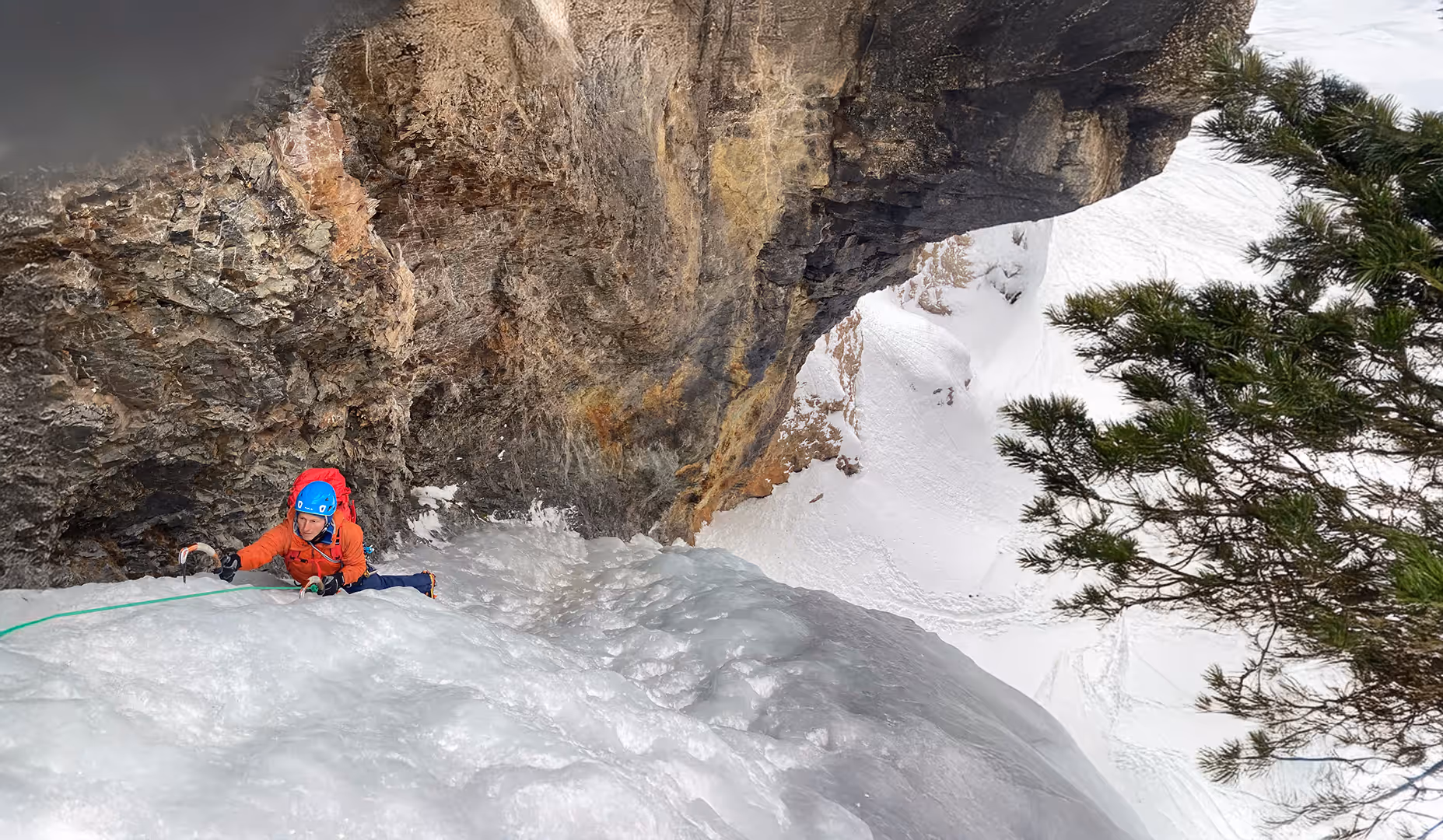 A climber in red gear ascends a steep icy slope near a rocky cliff face.