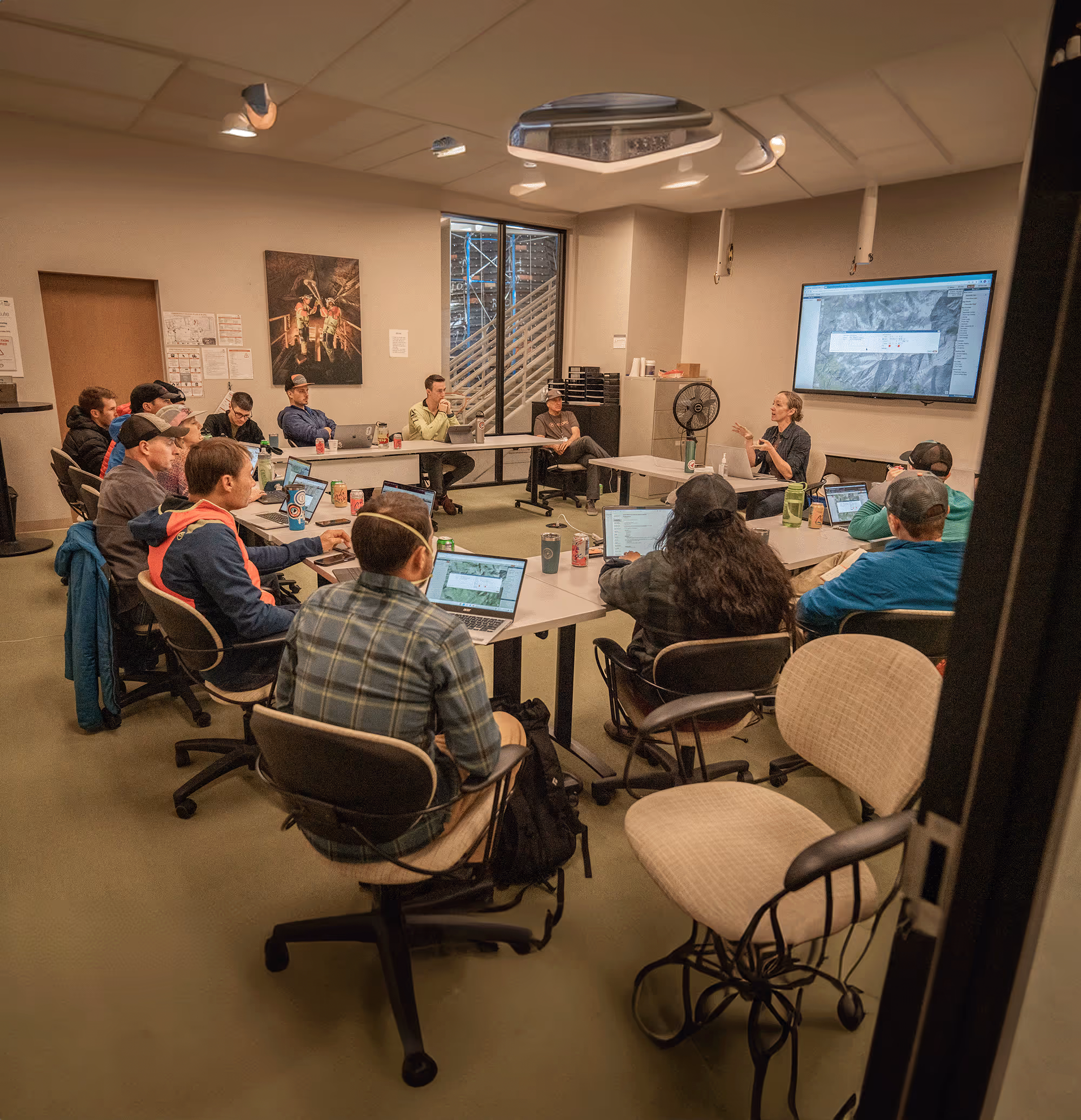 A group of people sit at a large table in a meeting room, looking at a screen.