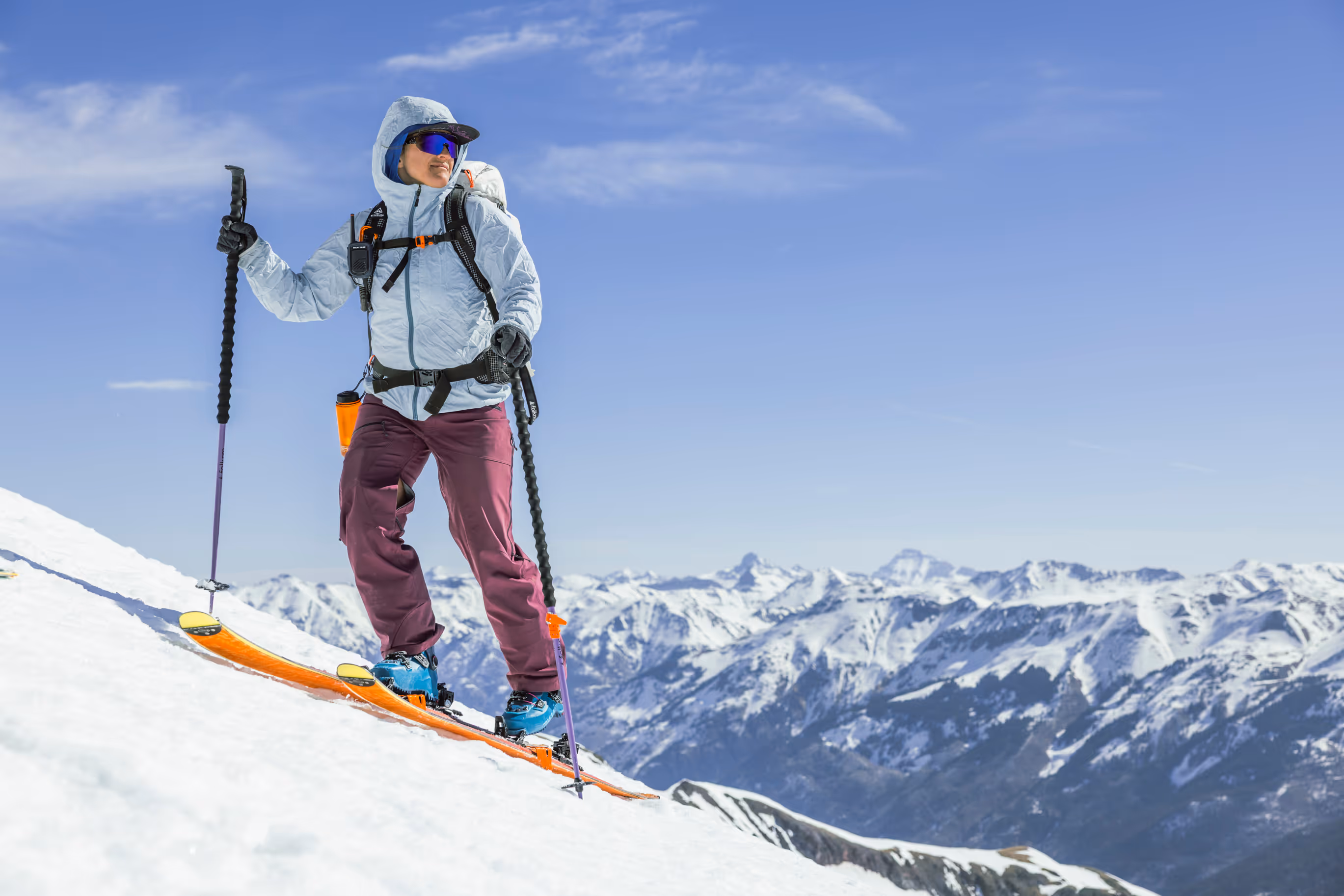 Person wearing ski gear and sunglasses stands on snowy mountain slope with poles, surrounded by snow-covered peaks.