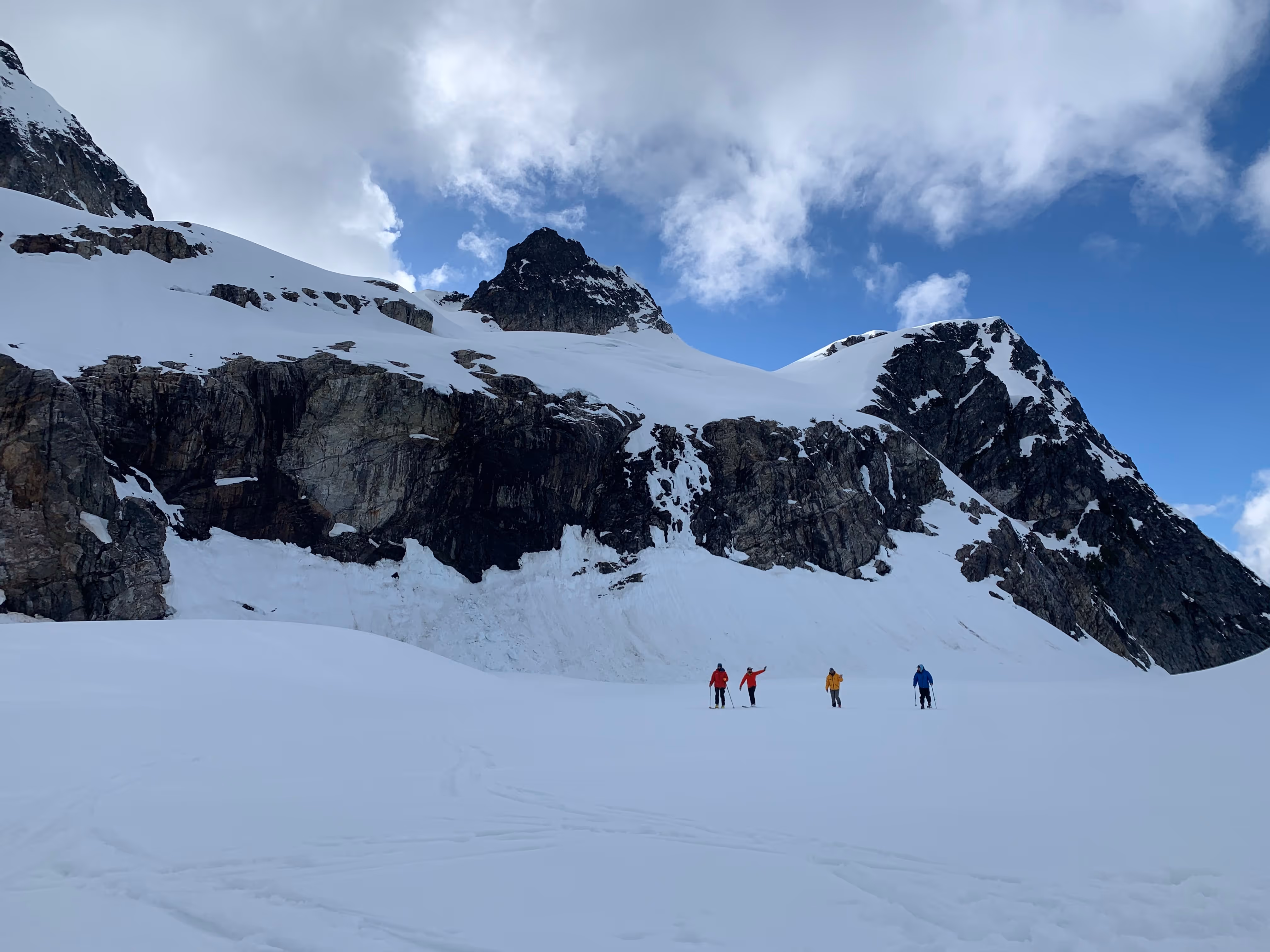 Four people in colorful jackets hike together on a snowy mountain under a partly cloudy sky.