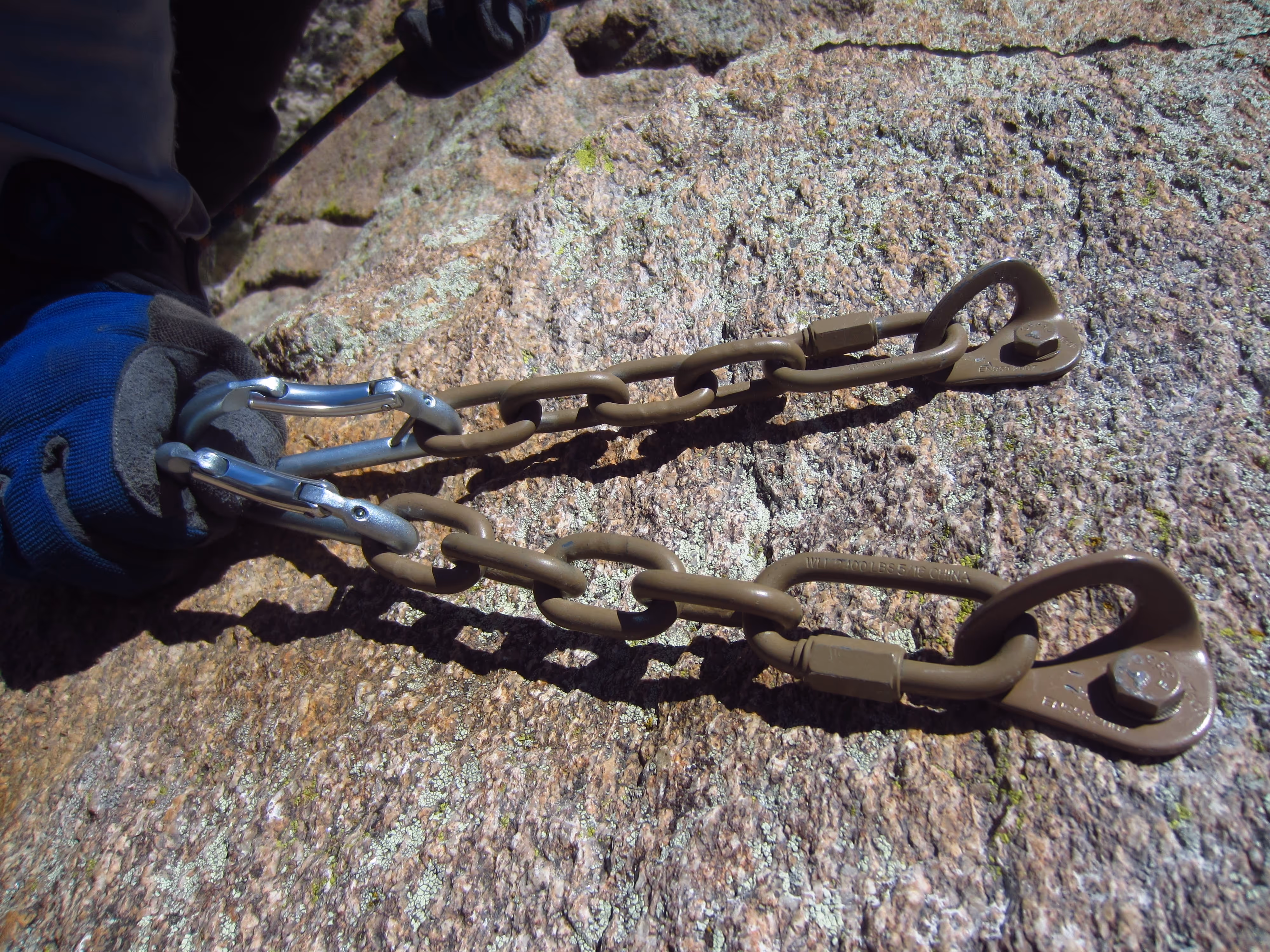 A person is attaching blue-gloved hands to metal chains secured to a rock surface with carabiners.
