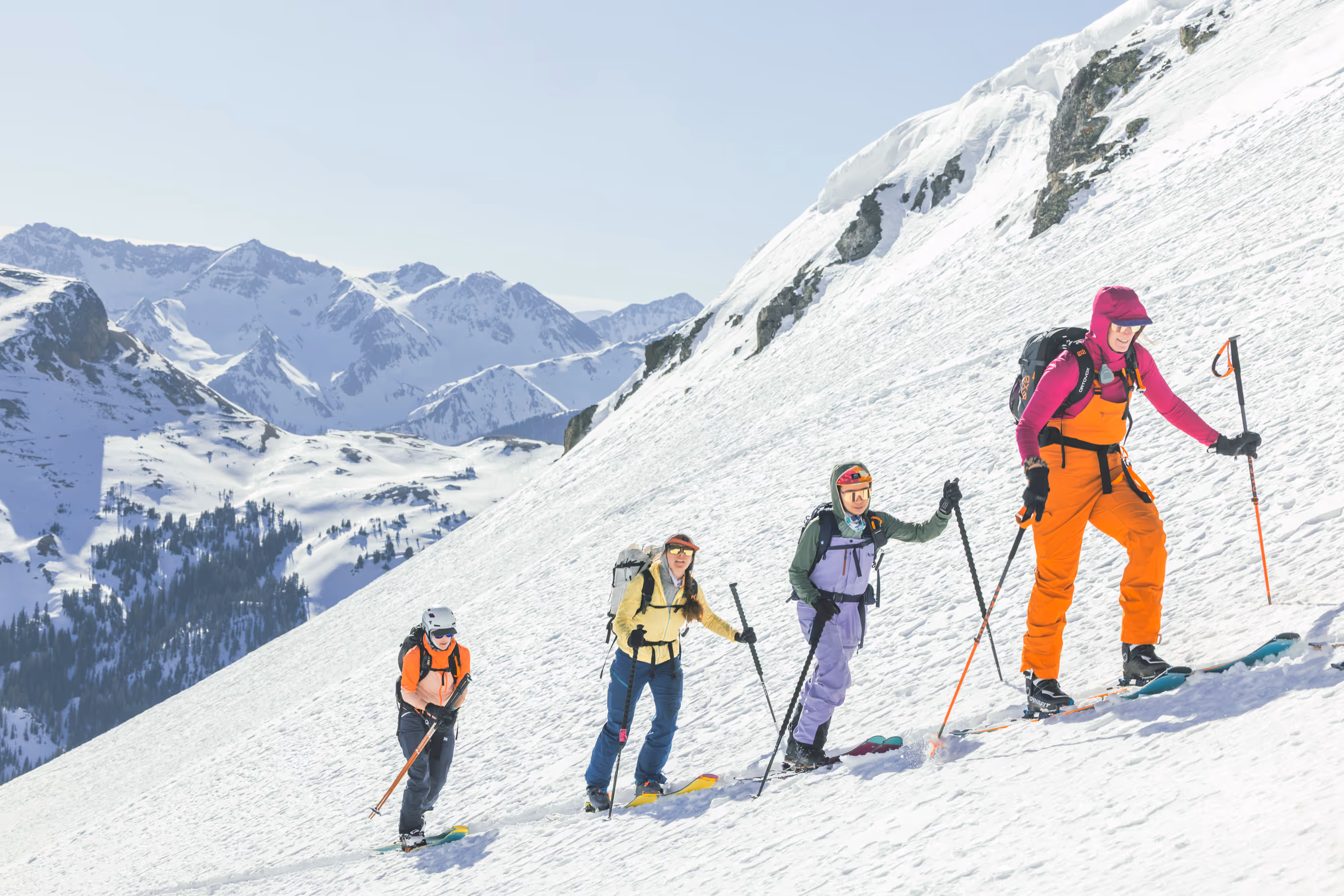 Four people in colorful gear are hiking up a snowy mountain with skis and poles.