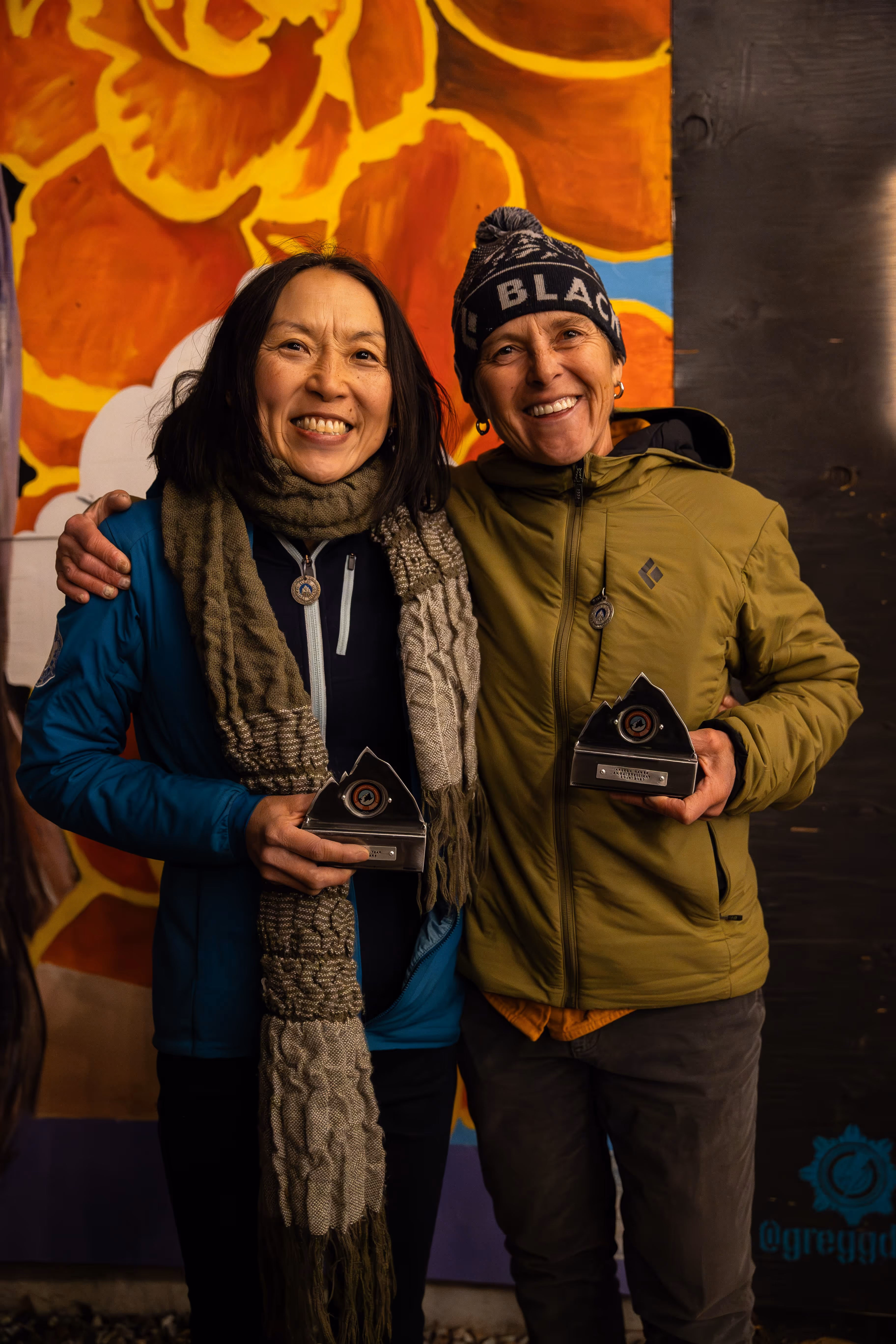 Two people smiling and holding trophies, standing in front of a colorful backdrop.