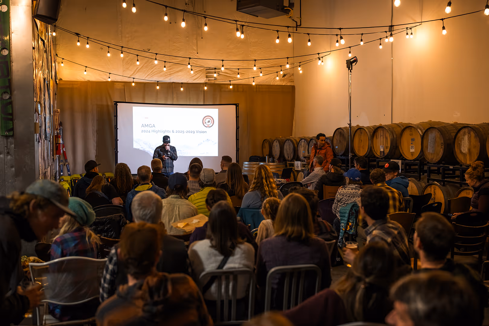 Person giving a presentation to an audience in a room with barrels and string lights.