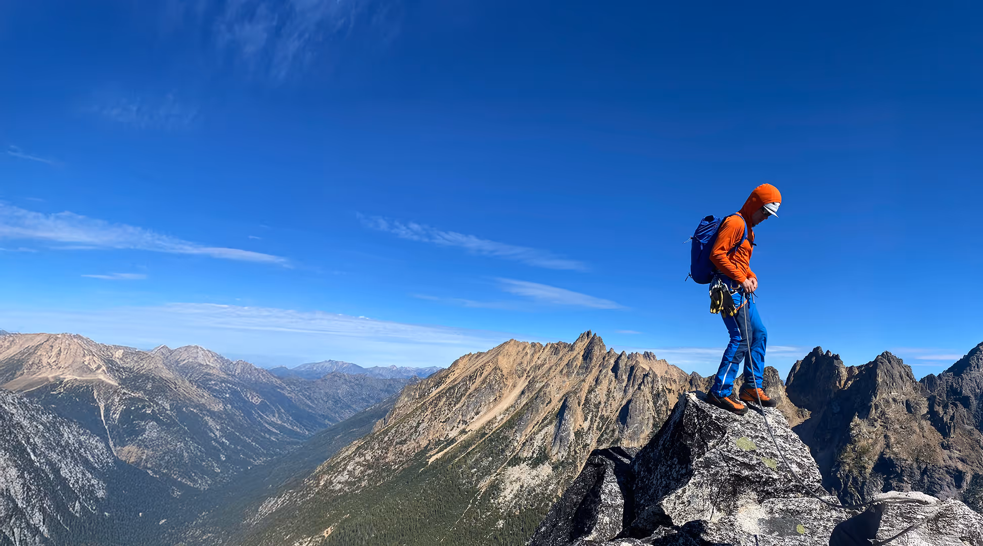 A person in hiking gear stands on a rocky peak with mountains in the background under a clear sky.