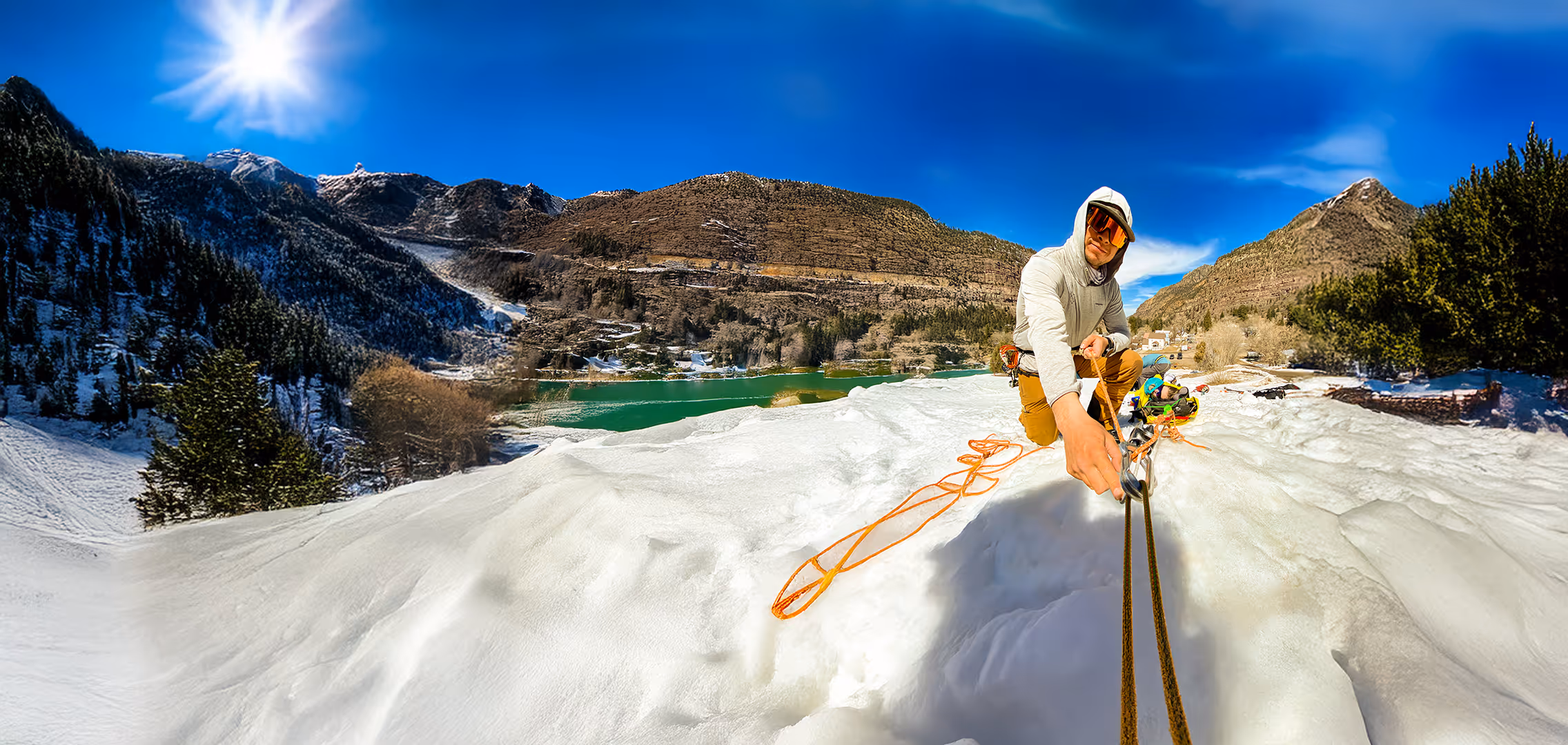 A person wearing a hoodie climbs a snowy mountain using ropes under a bright blue sky.