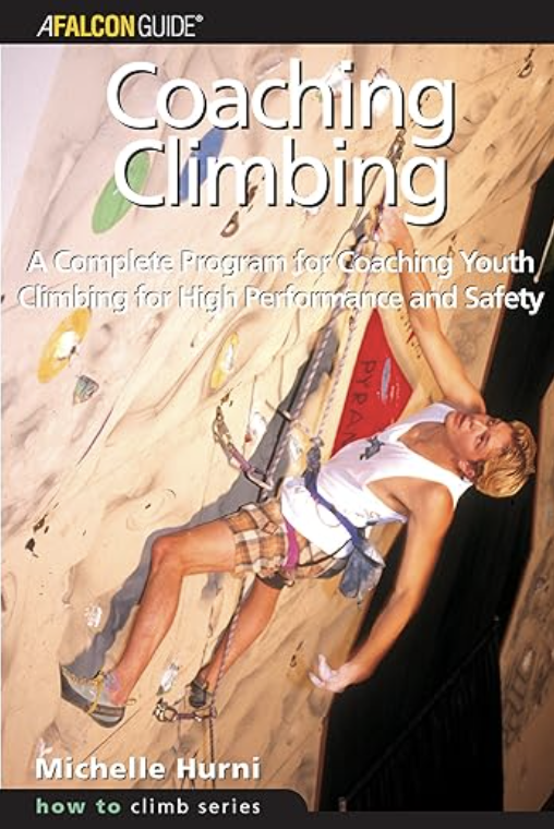 Person climbing an indoor rock wall with colorful handholds, wearing a white tank top and harness.