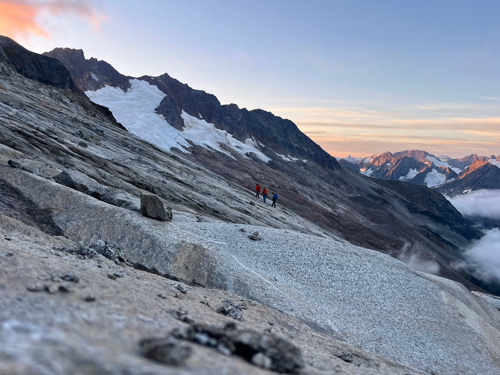 Three people hike on a rocky mountain slope with snow and distant peaks visible.