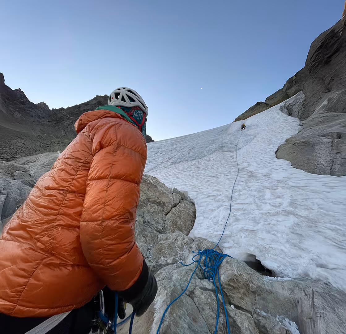 A climber in an orange jacket watches another person ascending a snowy mountain slope.
