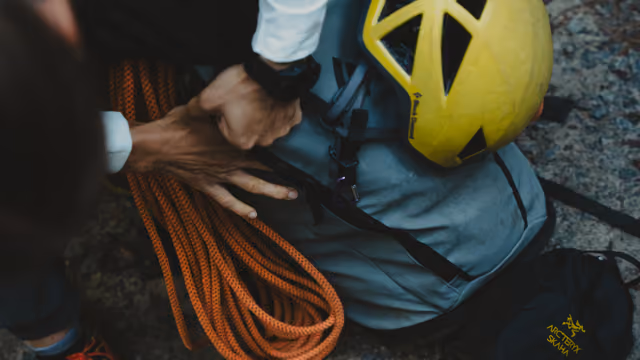 Person preparing climbing gear with an orange rope, blue backpack, and yellow helmet on rocky ground, adjusting straps.