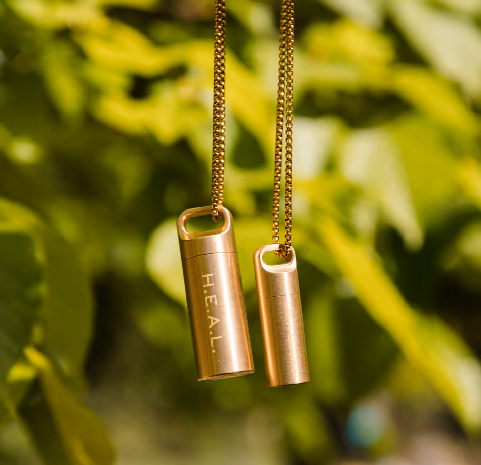 Two rose gold cylindrical pendant necklaces with 'HEAL' text, suspended on gold chains against green foliage background