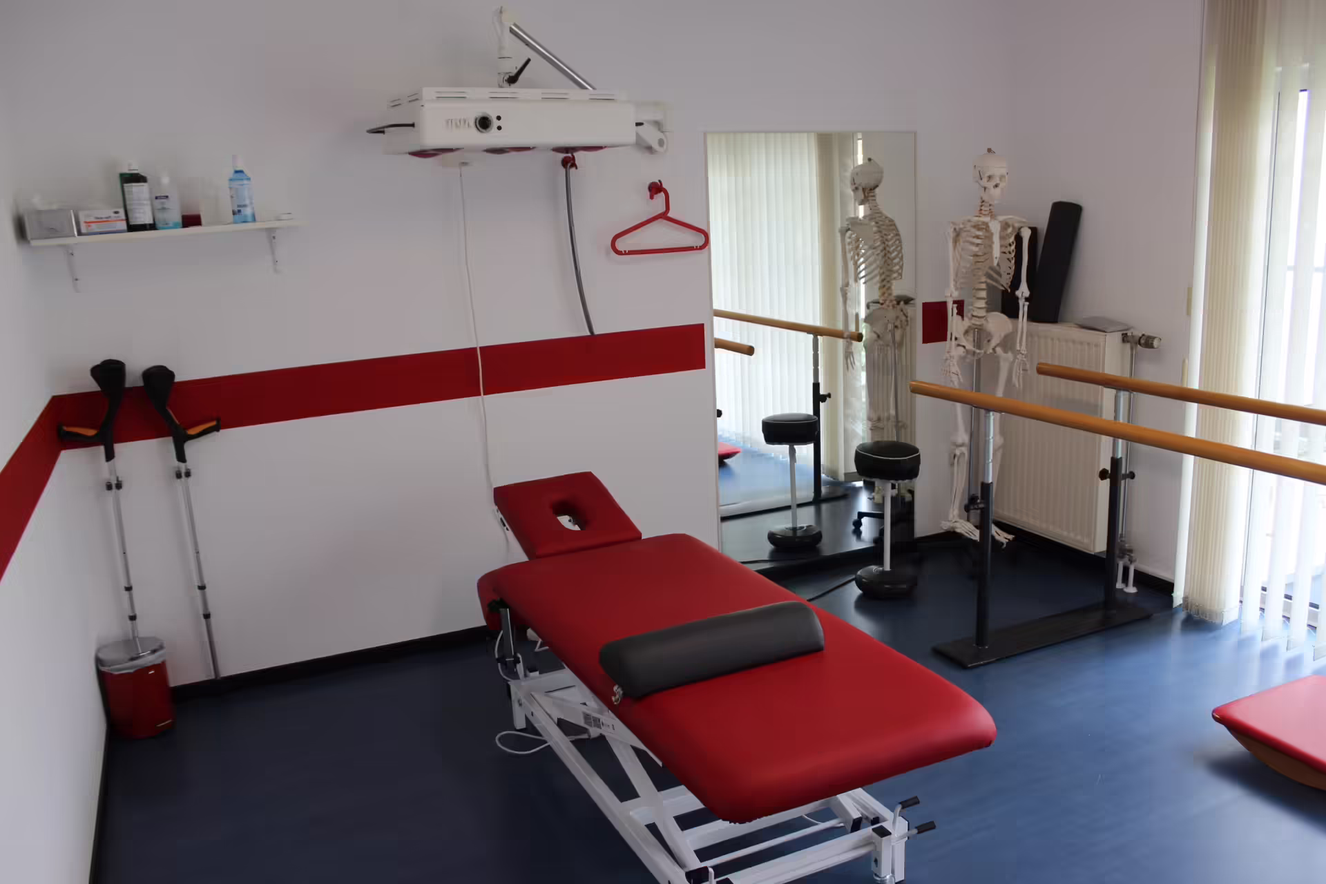 Physiotherapieraum mit roter Behandlungsliege, Spiegel, Übungsbarren, zwei Skelettmodellen, Krücken und medizinischen Utensilien an der Wand.