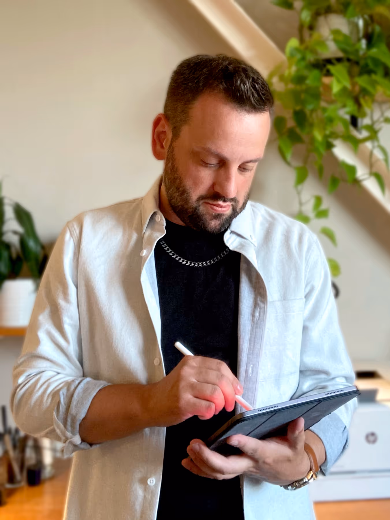 Man with short dark hair and beard using a stylus on a tablet in a room with plants and shelves.