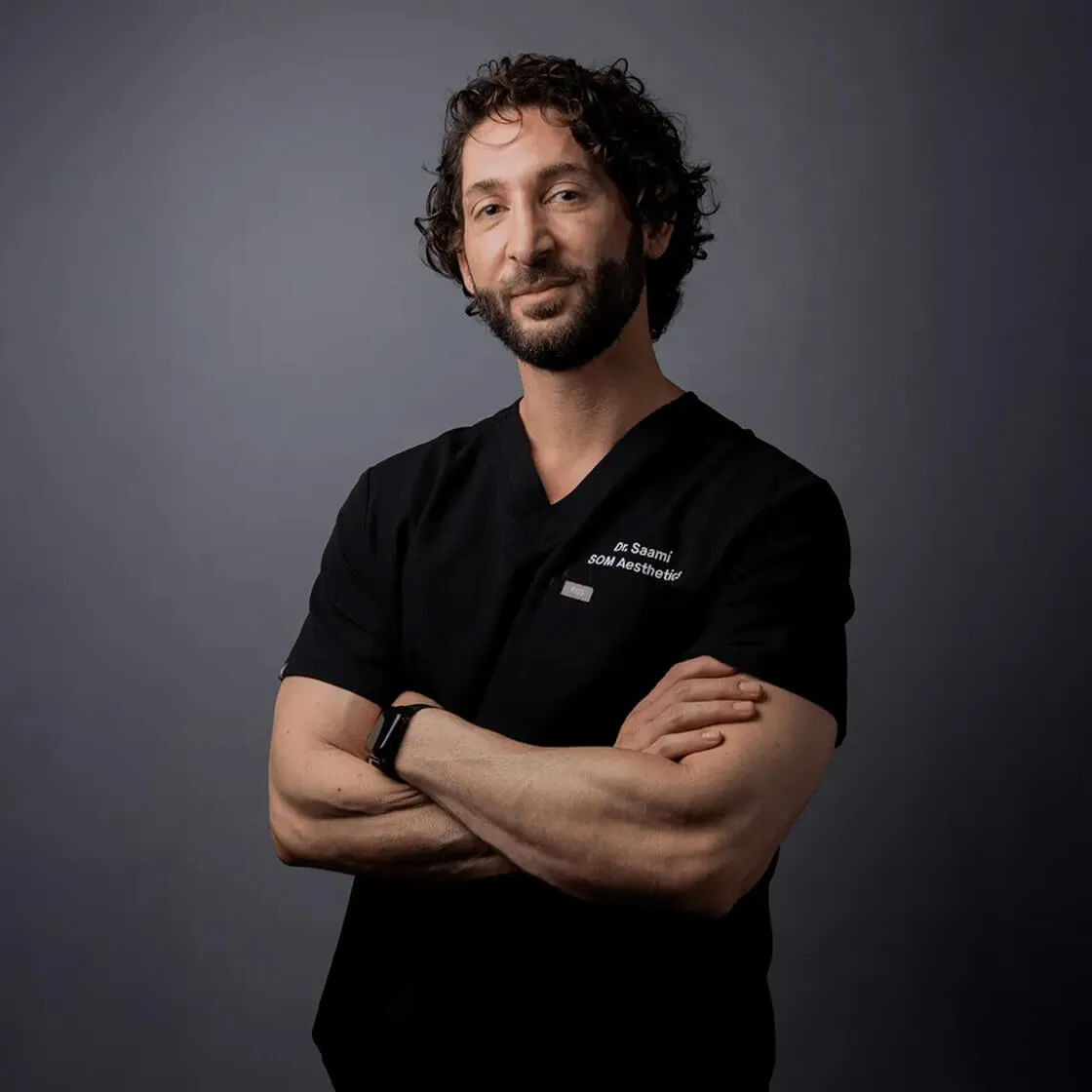 Man with curly hair and beard wearing black scrubs with text 'Dr Saami SOM Aesthetics', arms crossed, against a gray background.