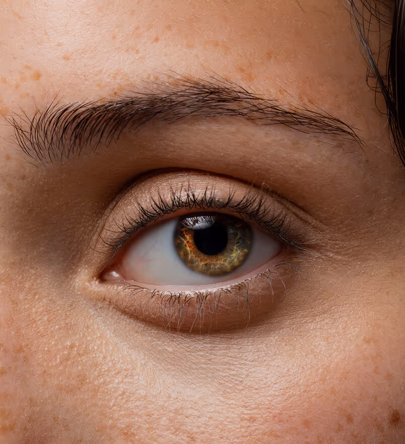 Close-up of a woman's face with clear, smooth skin, blue-gray eyes, and lightly touching her cheek with tattooed fingers wearing a gold ring.