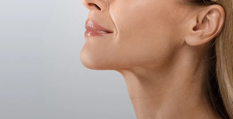 Close-up of a woman with eyes closed receiving a facial skincare treatment with cream applied on her cheek and a hand gently touching her face.