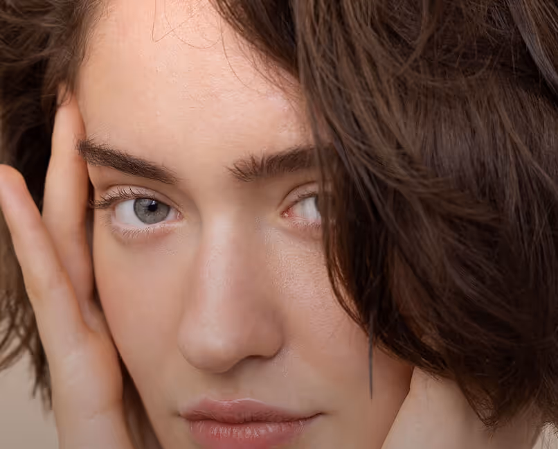 Close-up of a woman with blue eyes touching her face, wearing two rings on her fingers.