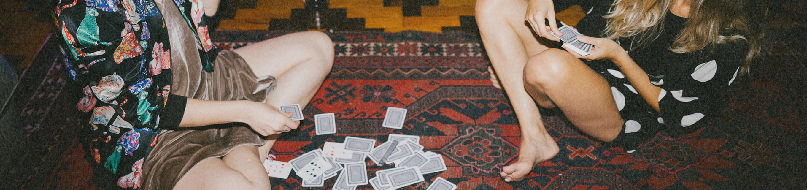 Two people sitting cross-legged on a patterned rug playing cards with a pile of cards between them.