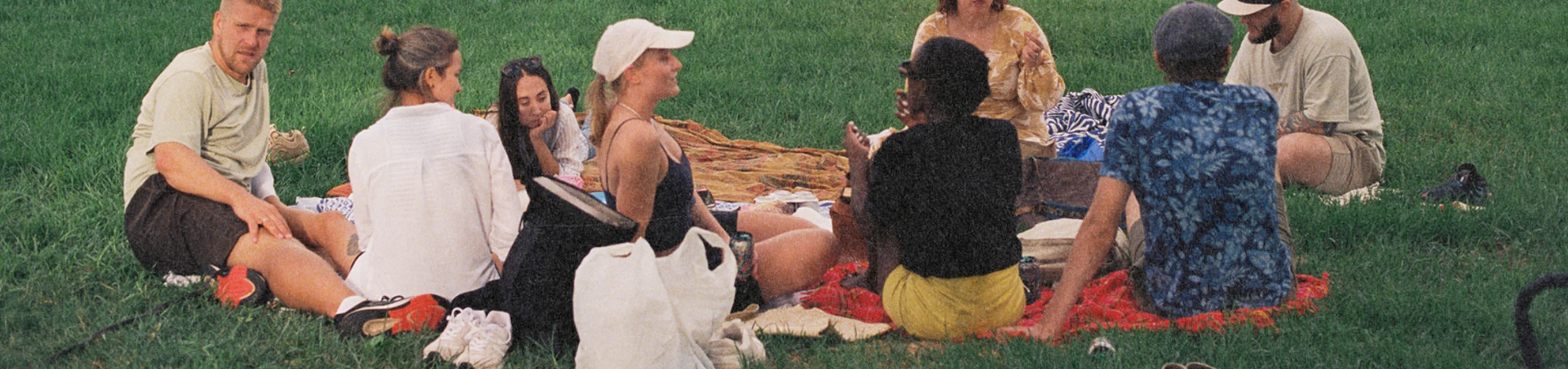 Group of seven people sitting on blankets having a picnic on green grass outdoors.