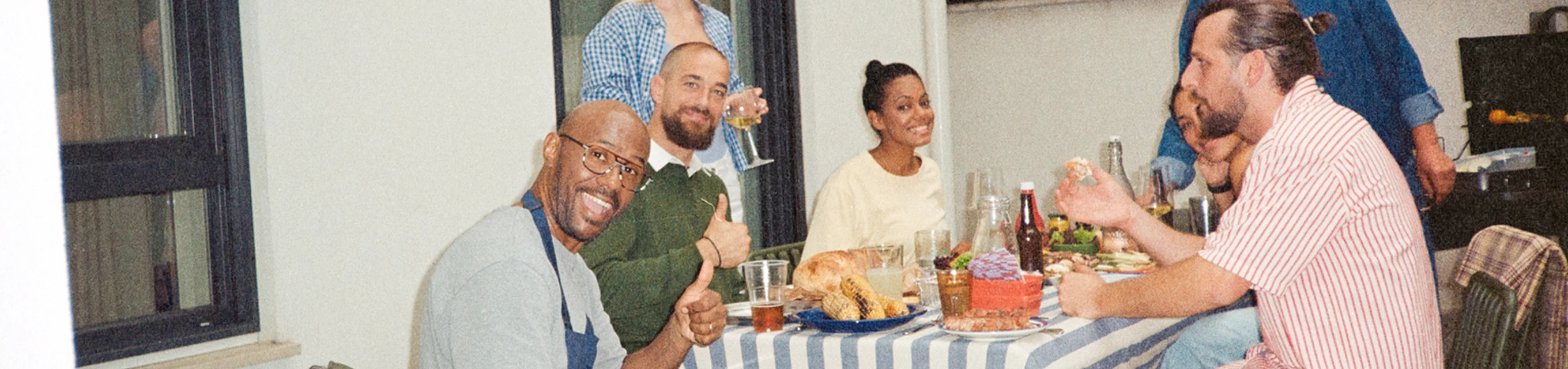 Five friends sitting at a table with food and drinks, smiling and giving thumbs up indoors.