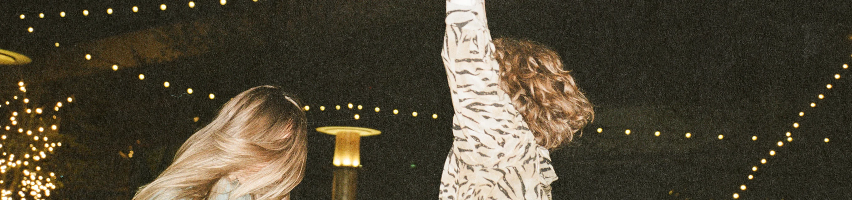 Two women dancing outdoors at night under string lights, one with arm raised and curly hair.