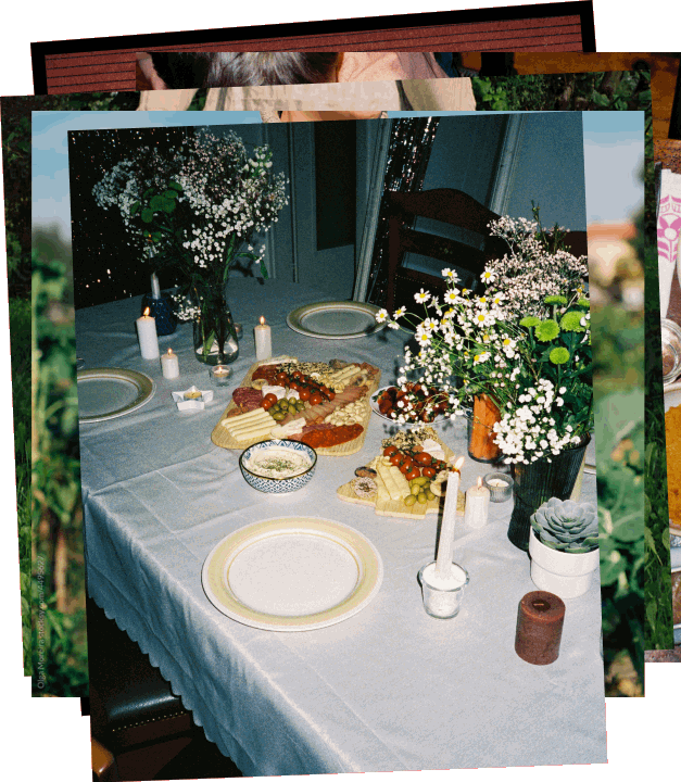 Dining table with white tablecloth set with empty plates, a cheese and vegetable platter, bowls of dip, candles, and floral arrangements including daisies and baby's breath.