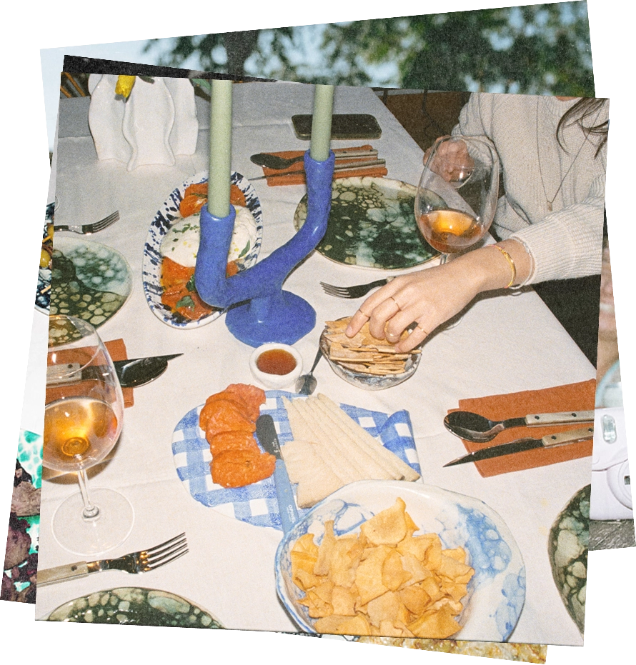 A dining table set with plates, cutlery, a glass of rosé wine, and a variety of food including sliced cheese, crackers, chips, and a dish with tomato slices. A person wearing a white sweater is reaching for crackers while holding a wine glass. A blue double-candle holder with green candles is at the center of the table.