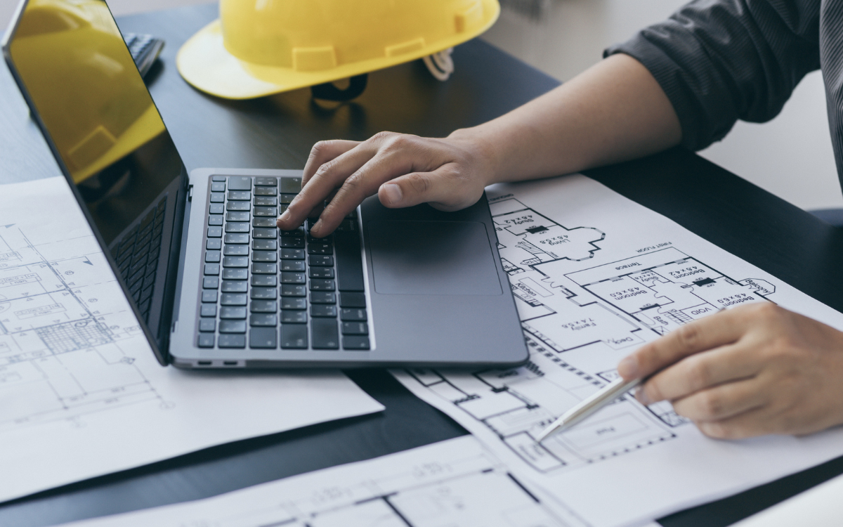 Close-up of a person working on a laptop with architectural blueprints and a yellow construction helmet on the table.