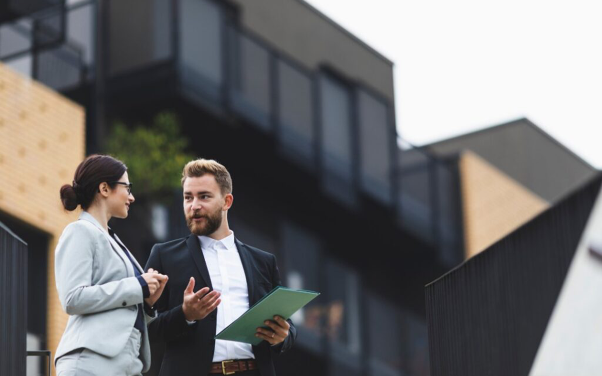 Two business professionals, a man and a woman, engaged in conversation outdoors near modern buildings.