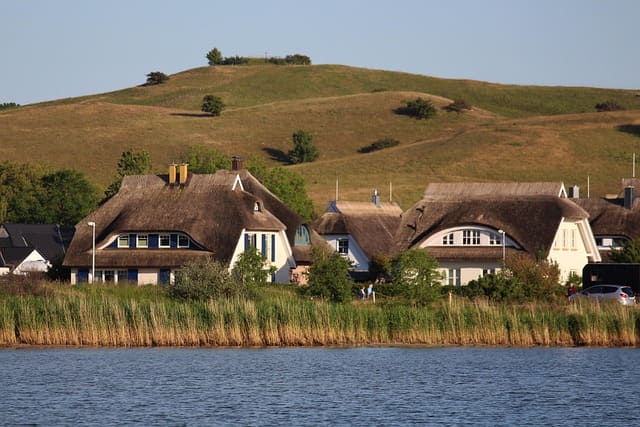 Traditionelle Reetdachhäuser am Ufer mit grünen Hügeln im Hintergrund.