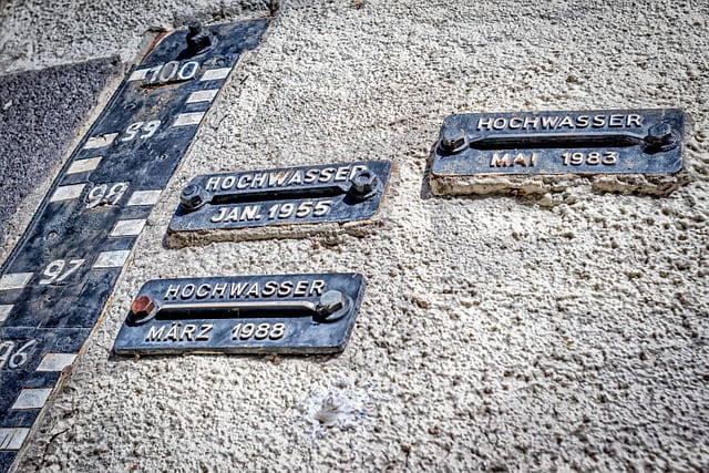 Mehrere Metallplaketten mit Jahresangaben früherer Hochwasser (1955, 1983, 1988) an einer Hauswand mit Pegelanzeige.