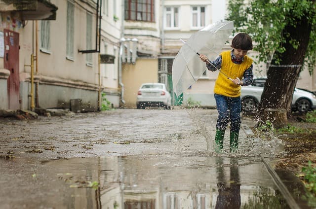Ein Kind mit gelber Weste und grünem Gummistiefeln springt in eine große Pfütze auf einer regennassen Straße.