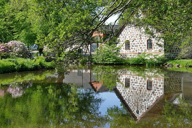 Naturstein-Gebäude am Ufer eines ruhigen Teichs mit Spiegelung im Wasser.
