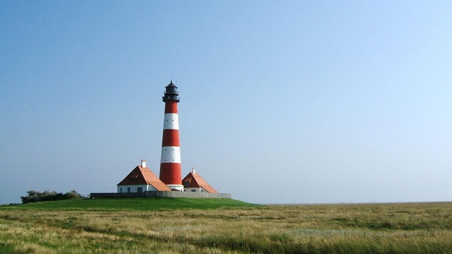 Der rot-weiß gestreifte Leuchtturm mit Nebengebäuden steht malerisch inmitten weiter Salzwiesen am Meer.