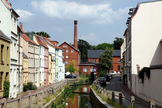Gasse mit farbigen Altstadthäusern entlang eines Kanals und einem alten Fabrikgebäude mit Schornstein im Hintergrund.