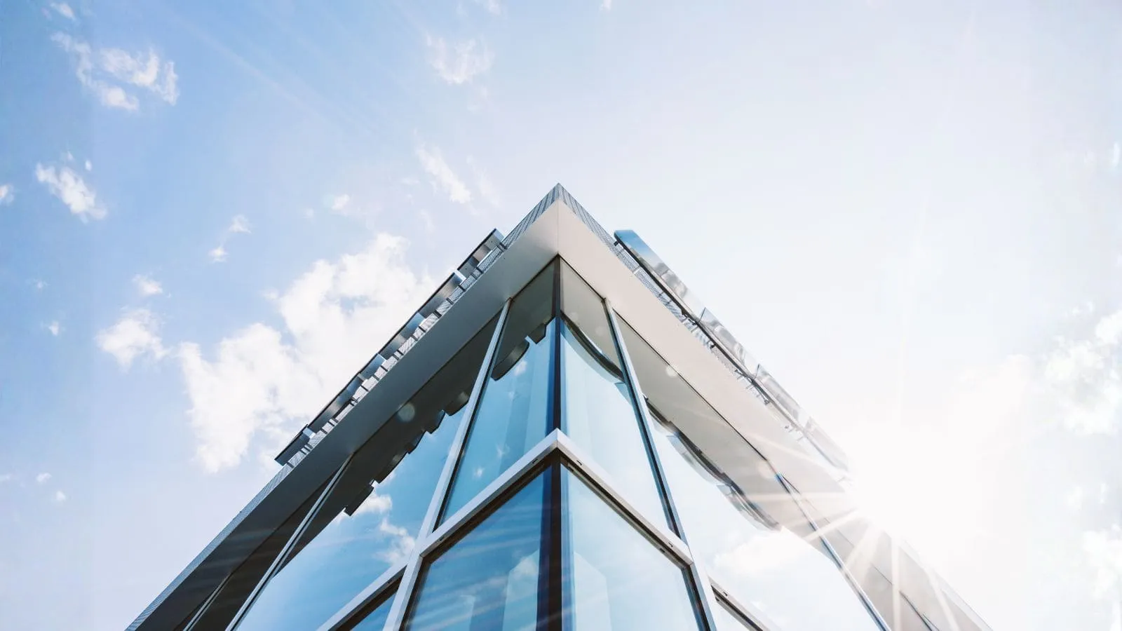 Low-angle view of a modern glass building corner with sun shining brightly and blue sky with clouds.