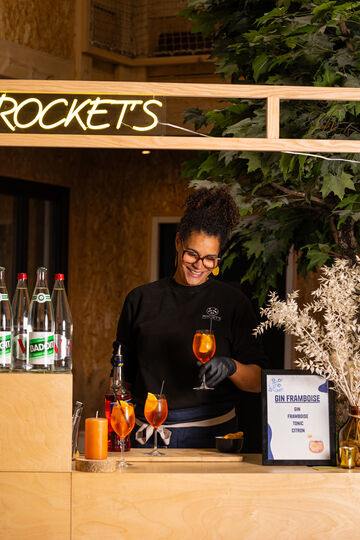 Serveuse souriante préparant un cocktail orange au bar sous un panneau lumineux «ROCKETS».