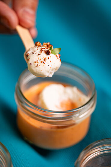 Cuillère en bois tenant une mousse blanche garnie d’épices au-dessus d'un pot en verre contenant un crémeux de tomates