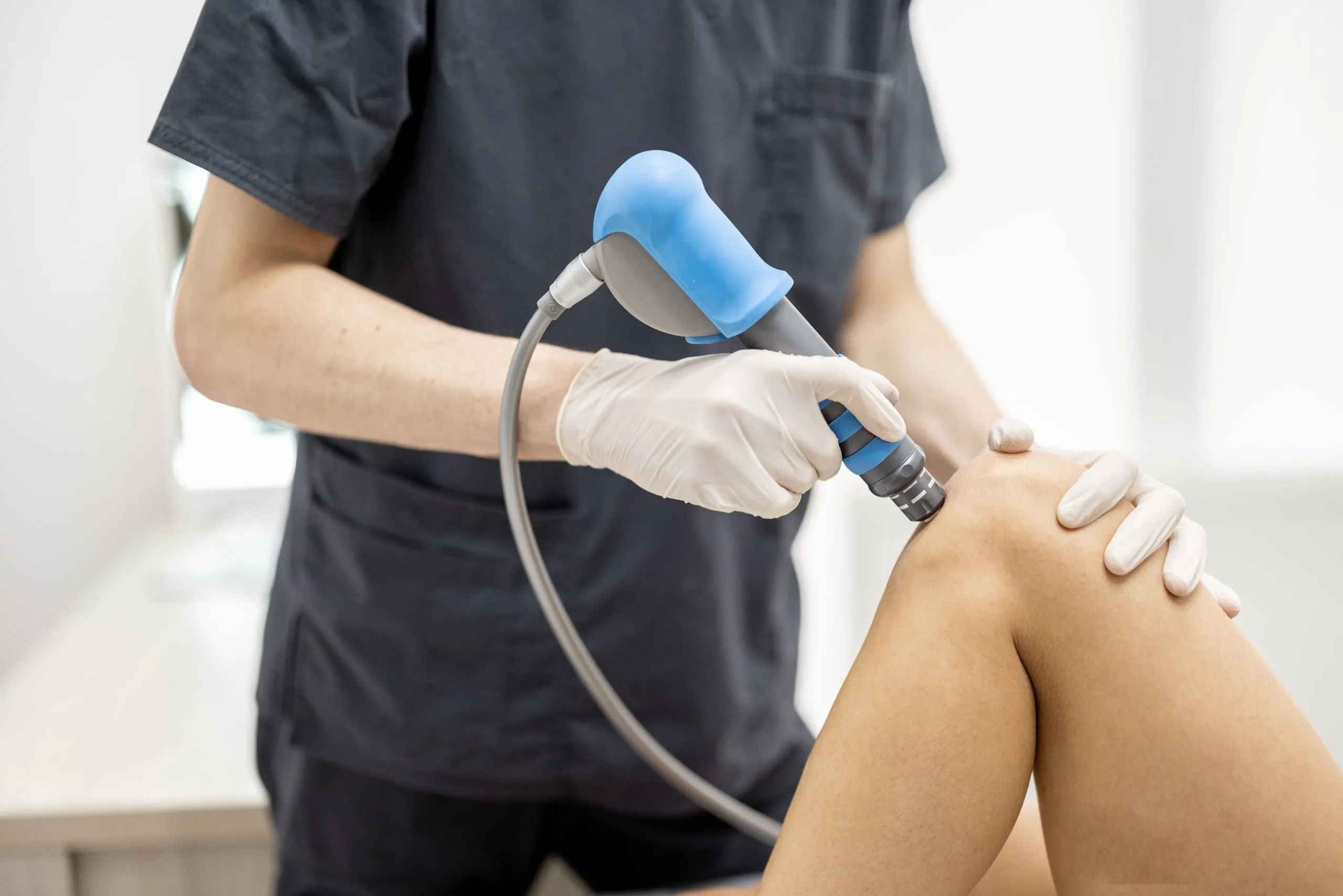 Medical professional using a shockwave therapy device on a bent knee of a patient.