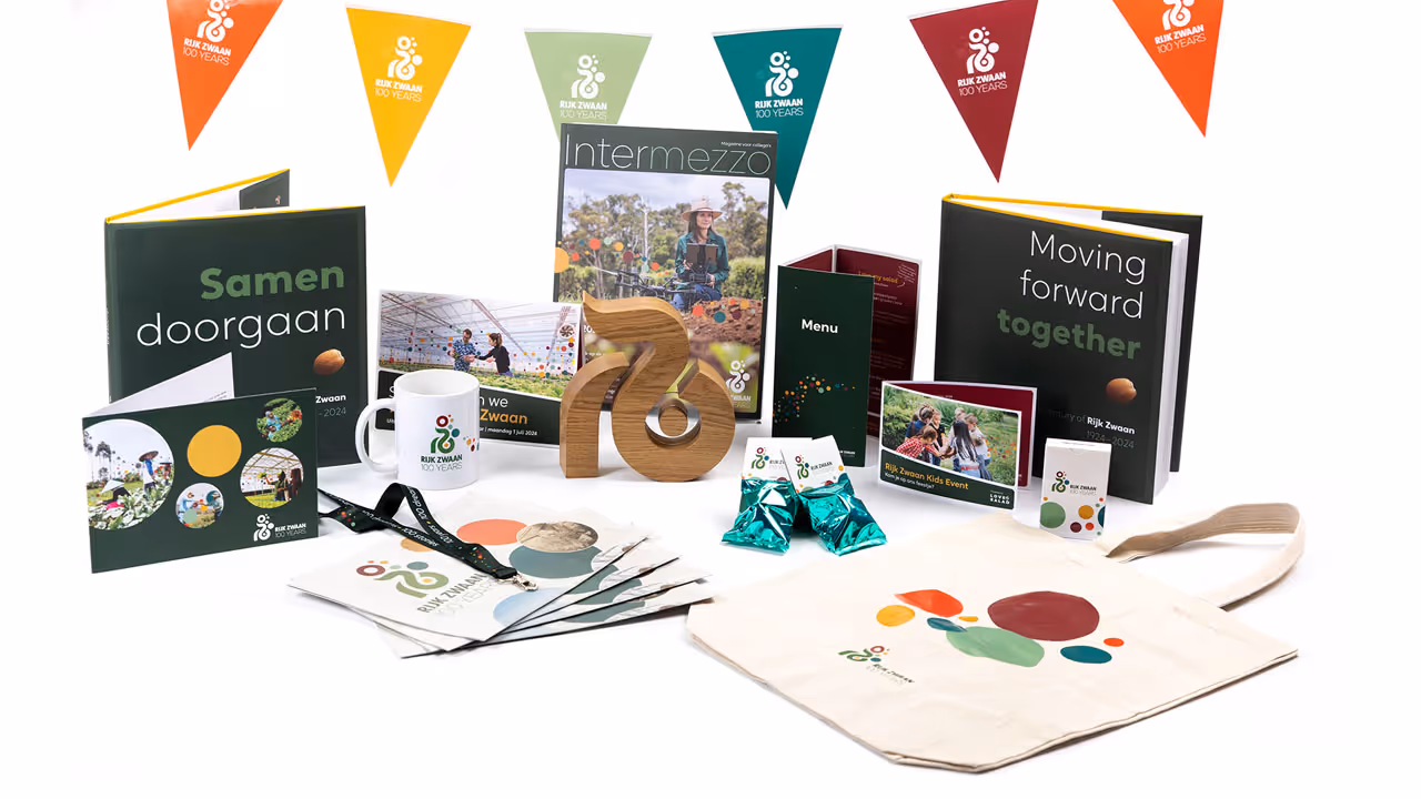 A collection of Rijk Zwaan anniversary items, including books, a mug, a wooden statue, flags, bag and leaflets, all with the 100-year logo and associated style elements created by the studio of campaign and event agency kaatenco