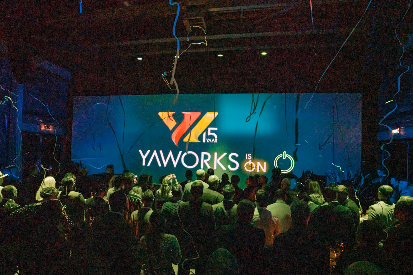 Kick-off evenement van de jaarcampagne YaWorks is On, met een groot scherm waarop het logo van Yaworks en de tekst '15 jaar Yaworks is on' te zien is, omgeven door feestelijke slingers. Georganiseerd door campagne- en evenementenbureau kaatenco