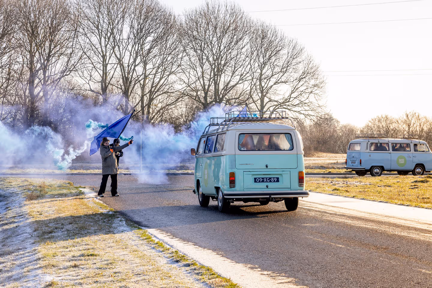 Two light blue Volkswagen vans drive on a road while two people wave a Vitam flag with blue smoke in an open landscape. The vans will be waved goodbye before the start of Vitam Goodcamp, organized by campaign and event agency kaatenco.