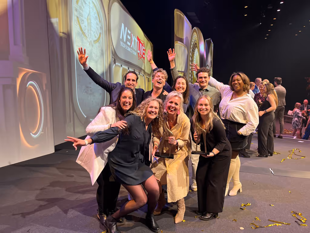 Blije groep van campagne- en evenementenbureau kaatenco die Gouden Giraffe award wint en poseert op het podium bij een evenement met een groot scherm en gouden confetti.
