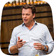 Carlo van Overveld als opdrachtgever van kaatenco en als man in een lichtblauw overhemd spreekt terwijl hij een pen vasthoudt, met een houten achtergrond.