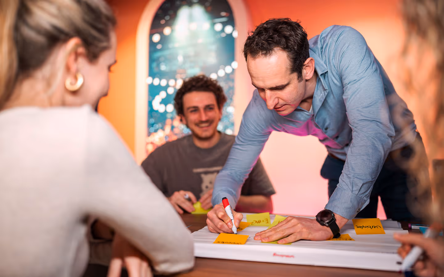 Luuk Drijfhout van Hooff als eigenaar van evenementenbureau kaatenco met een groep mensen werkt samen aan een project met notitiekaartjes op een tafel in een kleurrijke kamer.