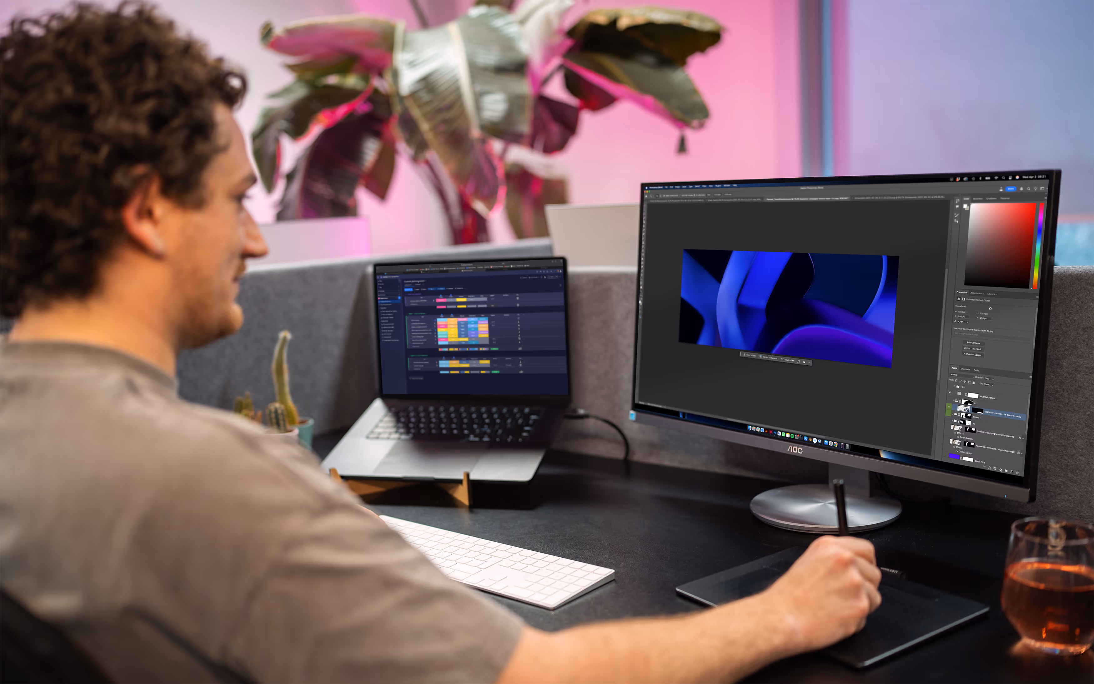 Man works with a drawing tablet at a desk with a large monitor and laptop in a modern office space with plants and pink mood lighting. The screen shows a planning tool and the other screen shows a design in Adobe.
