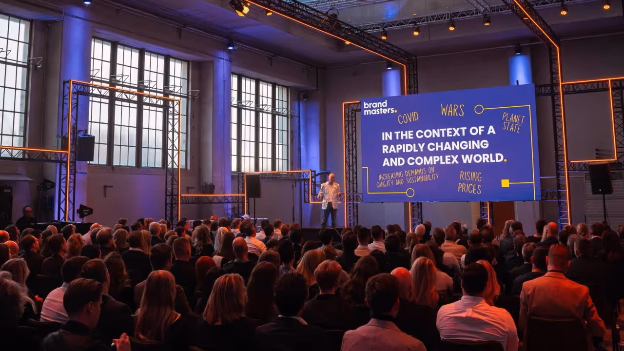 Een spreker staat op het podium tijdens het kick-off event voor Brand Masters. Hij staat voor een groot scherm met de tekst over een snel veranderende en complexe wereld, met een publiek dat luistert in een industriële evenementenzaal. Georganiseerd door campagne- en evenementenbureau kaatenco