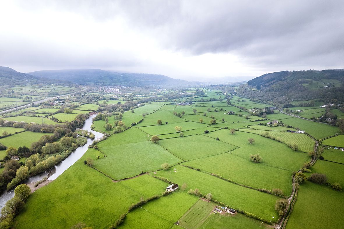 Aerial view of a river winding through a valley of green grassland fields and hedgerows under cloud